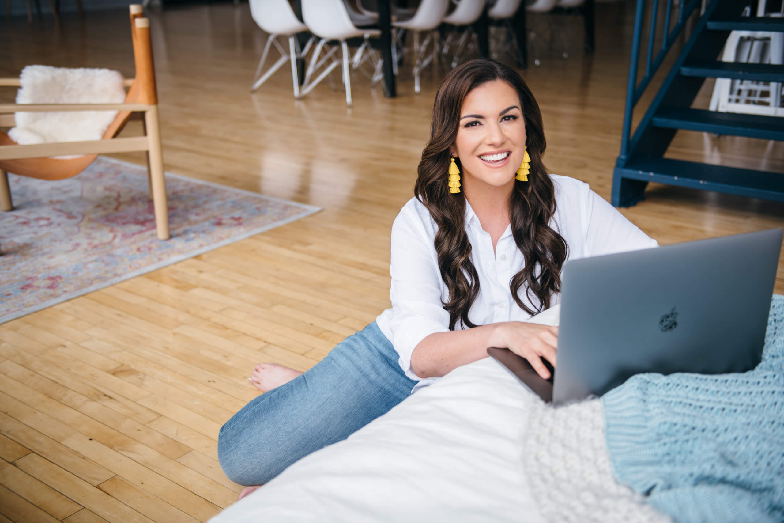 Amy Porterfield is sitting on the floor smiling at the camera with a laptop on the sofa in front of her.
