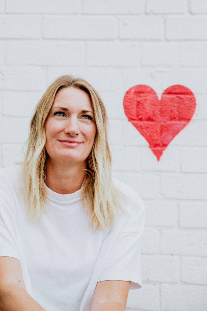 Marielle is wearing a white t-shirt and smiling. Behind her is a white wall with a red heard painted on it.